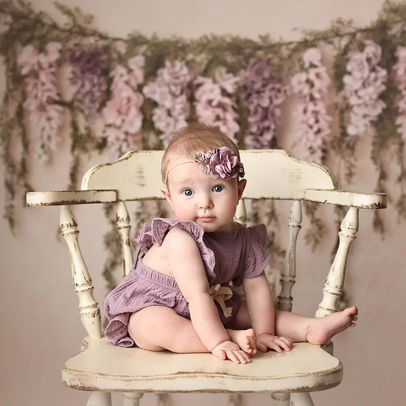 Kansas-city-sitter-session-portrait Baby girl sitter session in mauve romper, flower headband, sitting on vintage cream chair in front of a floral backdrop