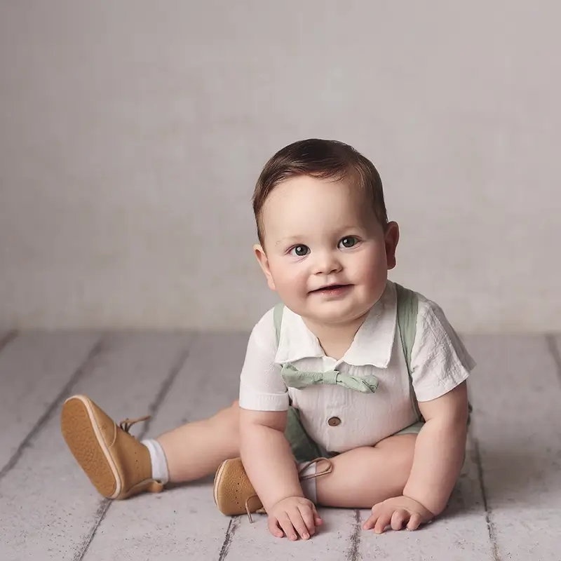 Sitter-Session-Portrait-Kansas-City Baby boy sitter portrait in white outfit with mint bow tie, smiling while sitting on the floor during studio photo session in Kansas City
