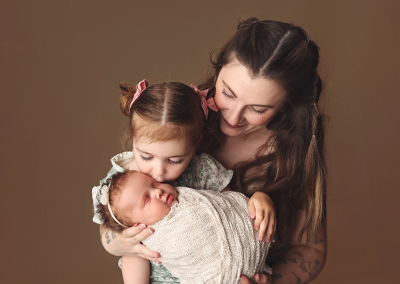 A new mom holding her toddler and newborn daughter on her lap while big sister kisses baby in Kansas City.