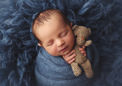 A KC newborn session of a baby boy wrapped in a blue fuzzy wrap and hugging a tiny teddy bear.