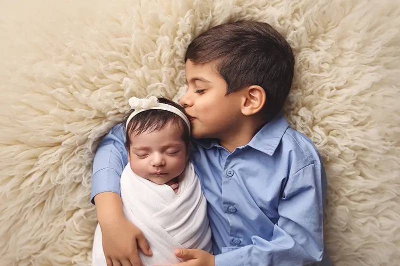 kansas-city-newborn-sibling-portrait Big brother kissing newborn sister during swaddled portrait on soft blanket at Blue Springs photography studio