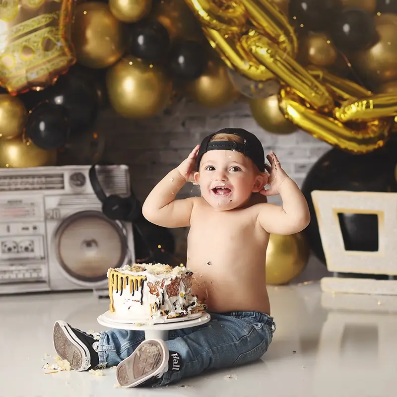 kansas-city-one-year-cake-smash-session One year old boy hip hop themed cake smash with gold balloon garland, boombox prop, baseball cap, and messy first birthday cake