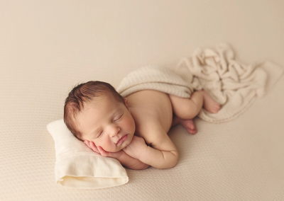 A newborn boy lays on a white cream blanket with a matching blanket draped over him while sleeping sweetly at his KC newborn photography session.