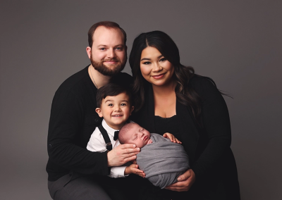 A family of four poses for a photo, toddler is helping to hold his newborn brother during his Kansas City Newborn Studio Session.