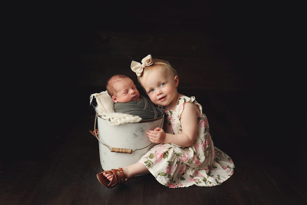 Big sister smiling while sitting beside newborn baby brother in cozy newborn portraits