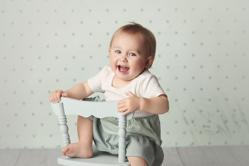 One-year-old girl smiling while sitting on tiny chair in professional portrait studio