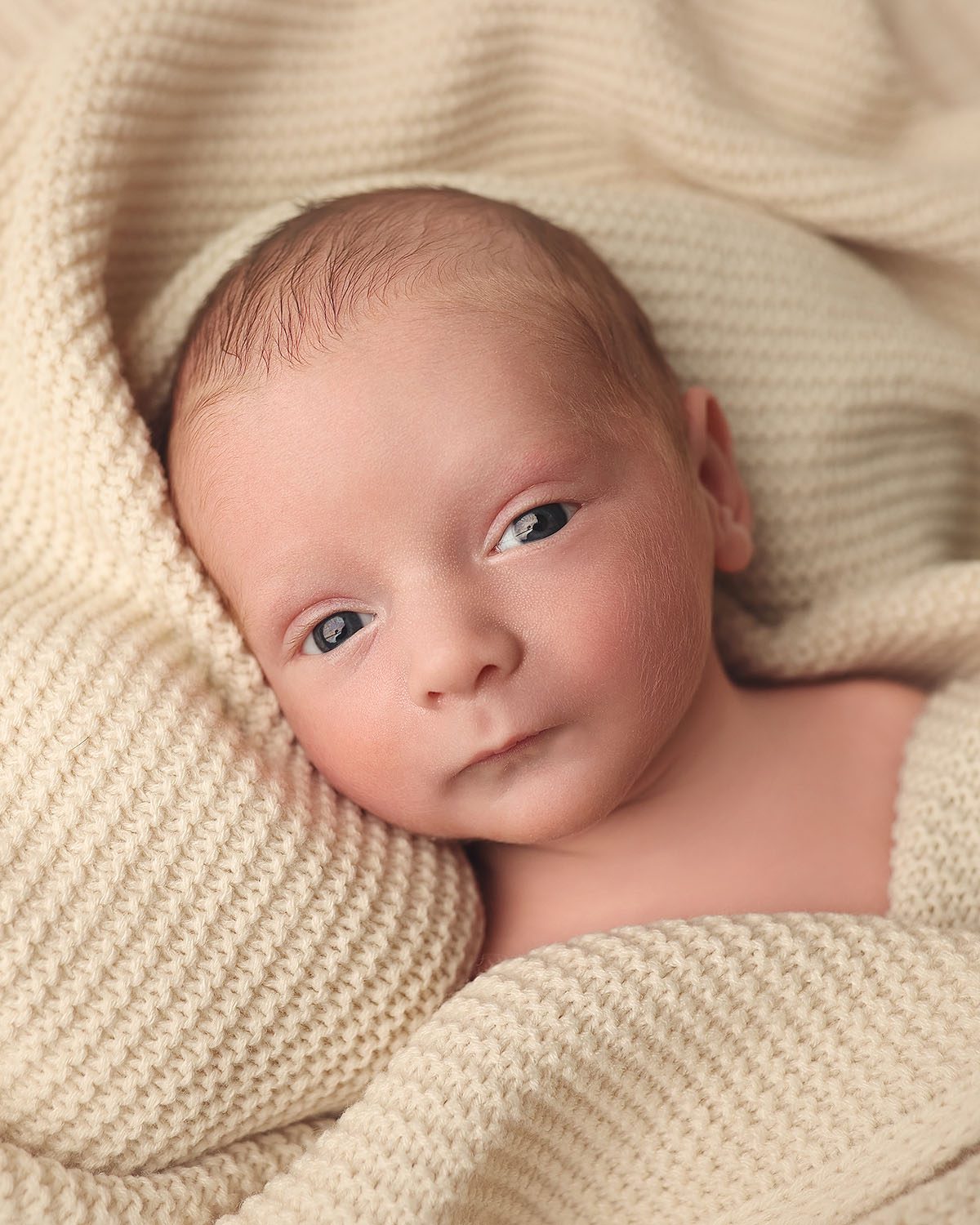 Close up portrait of a wide awake newborn baby boy looking toward the camera, wrapped loosely in a soft knit blanket