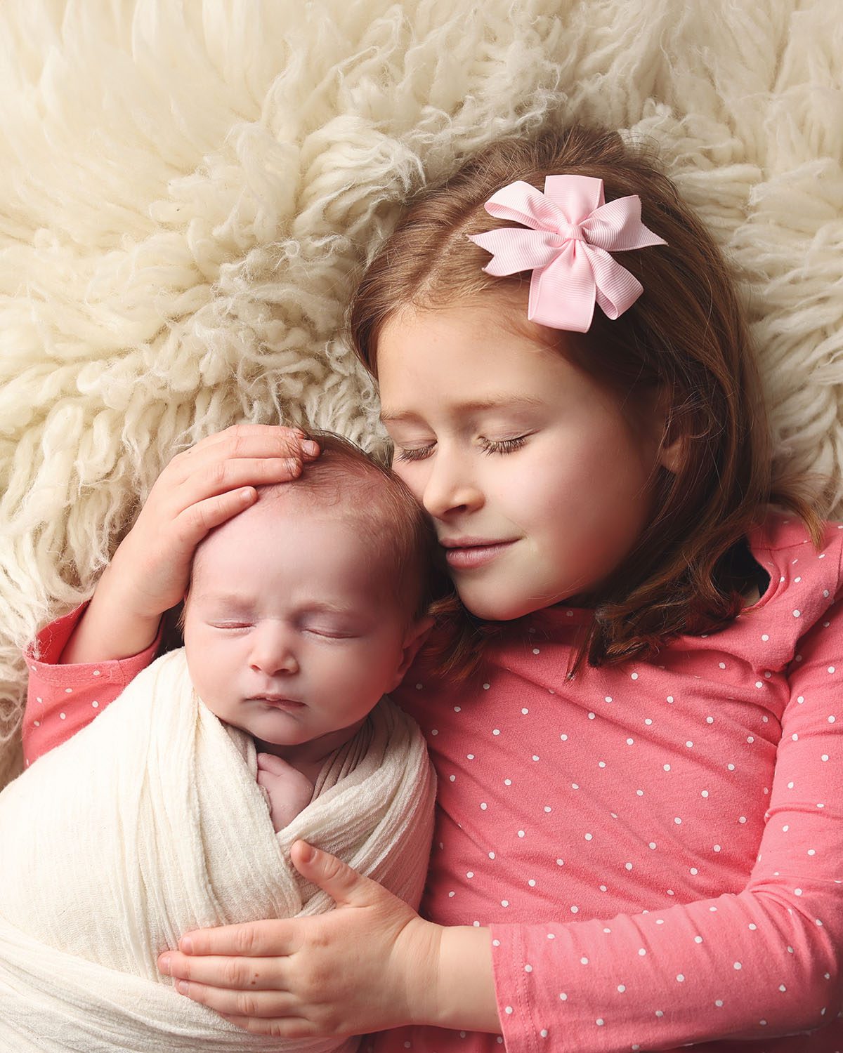 Sibling portrait with older sister holding her newborn baby brother, swaddled and resting together on a white rug during a newborn photography session.