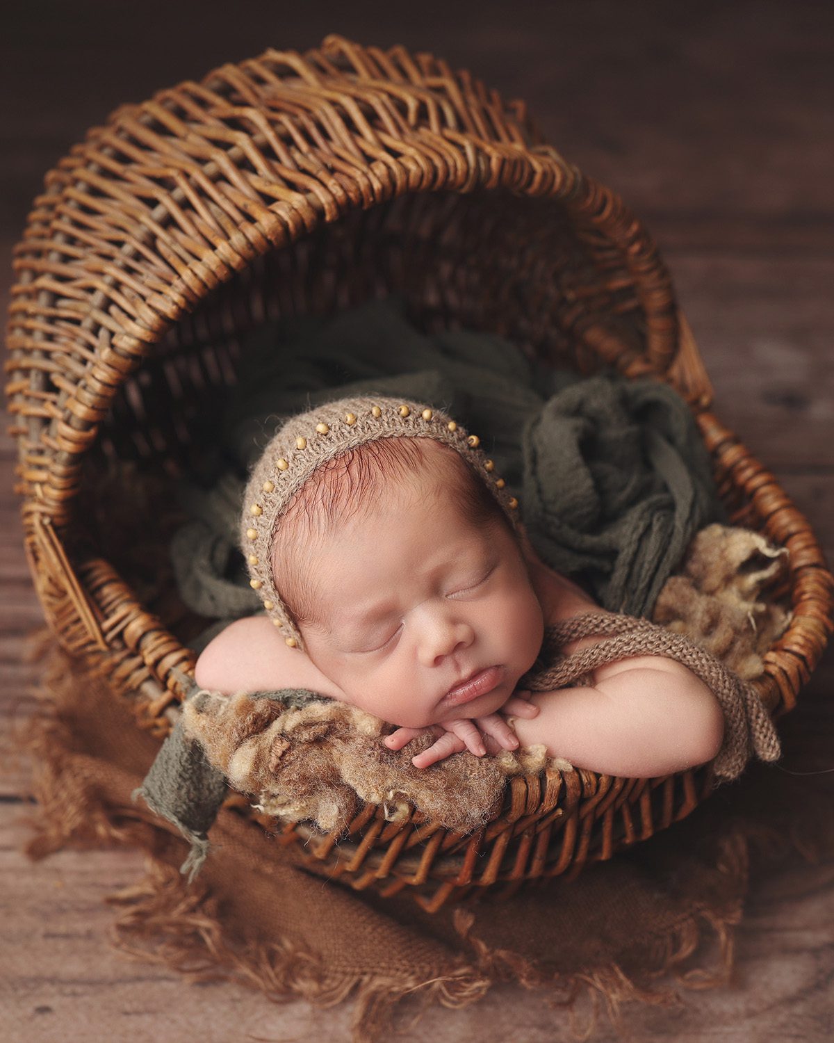 Newborn baby boy wearing a knit bonnet, posed in a wicker cradle basket during a studio newborn photography session.