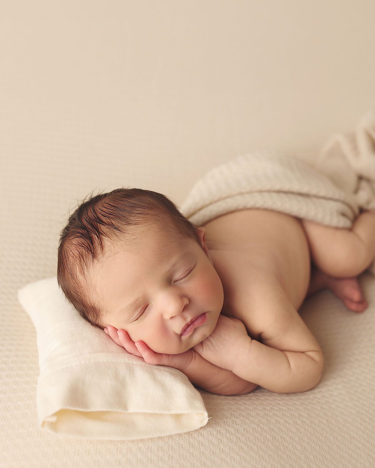 Newborn baby boy sleeping peacefully with his cheek resting on his hand, posed on a whit pillow with a textured blanket.