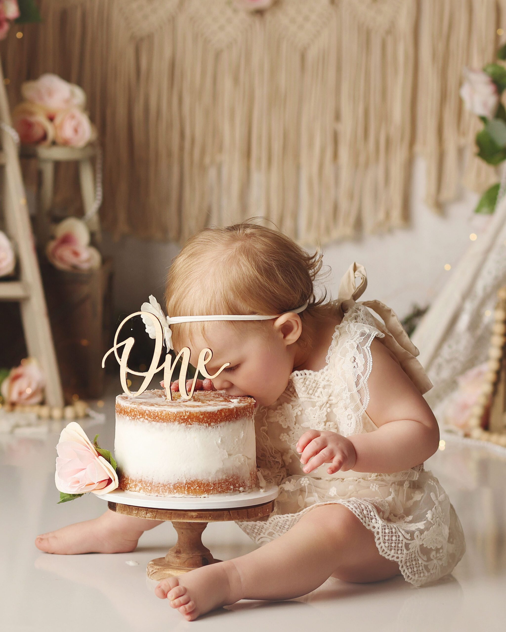 Boho style studio cake smash portrait of a one year old girl wearing a lace romper, captured leaning forward to take a bite of a simple cake during cake smash session