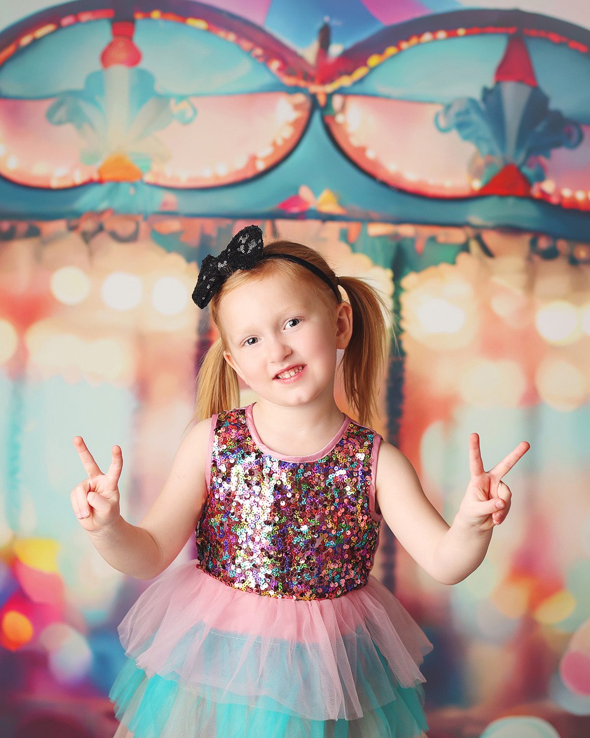Studio child birthday photo of a young girl wearing a sequin and tulle dress, posing with her hands in peace signs in front of a carousel backdrop.