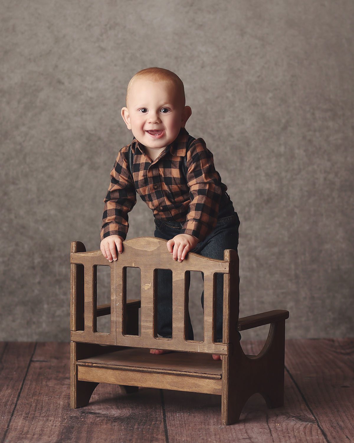 One year old boy milestone portrait wearing a plaid shirt, standing on a wooden bench and smiling at the camera