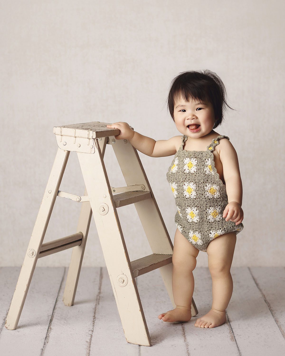 One year old girl milestone portrait in a crocheted romper holding onto a cream antique stepladder and smiling