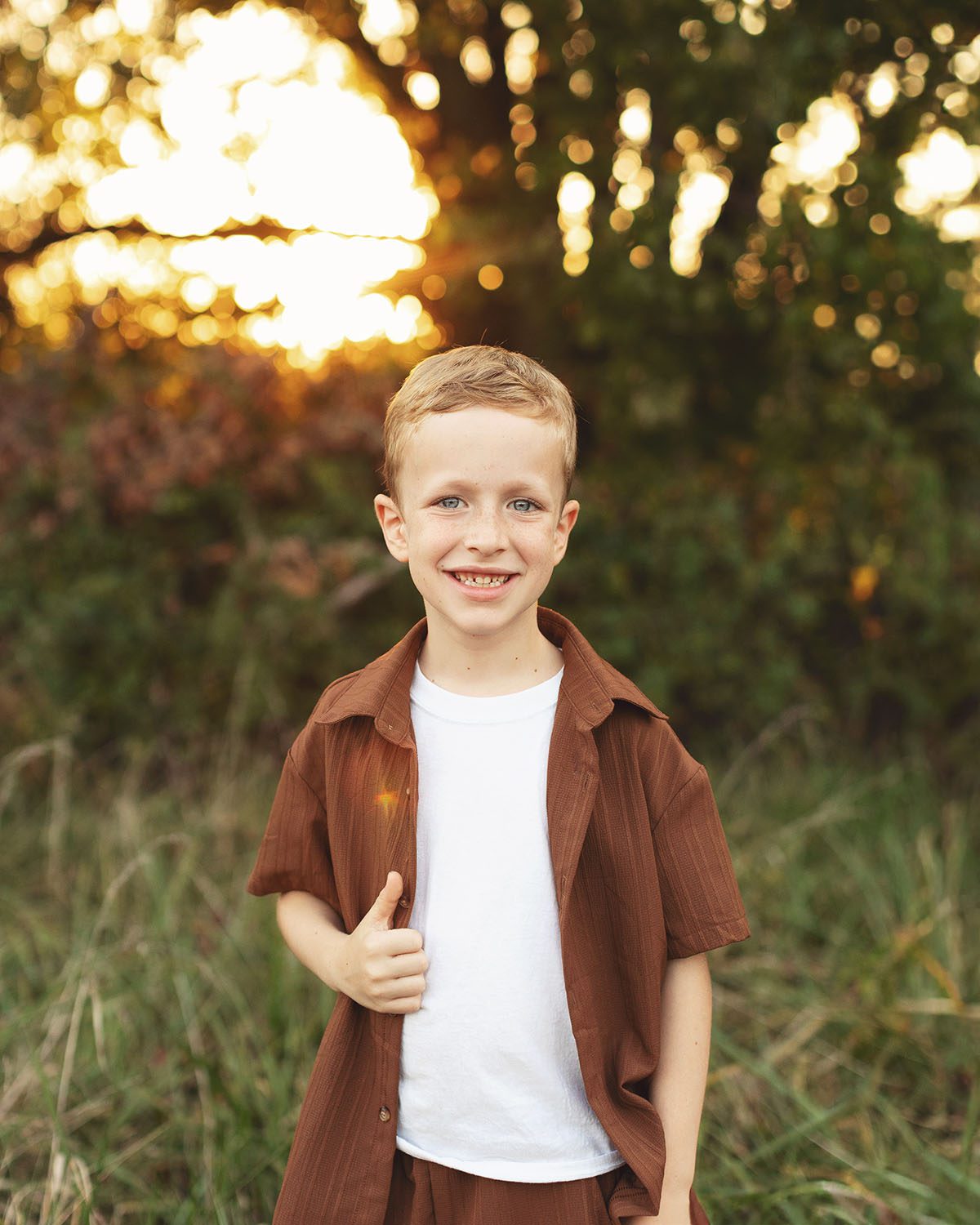 Outdoor child portrait at sunset featuring a smiling boy wearing a short sleeve linen shirt and shorts with freckles and blue eyes