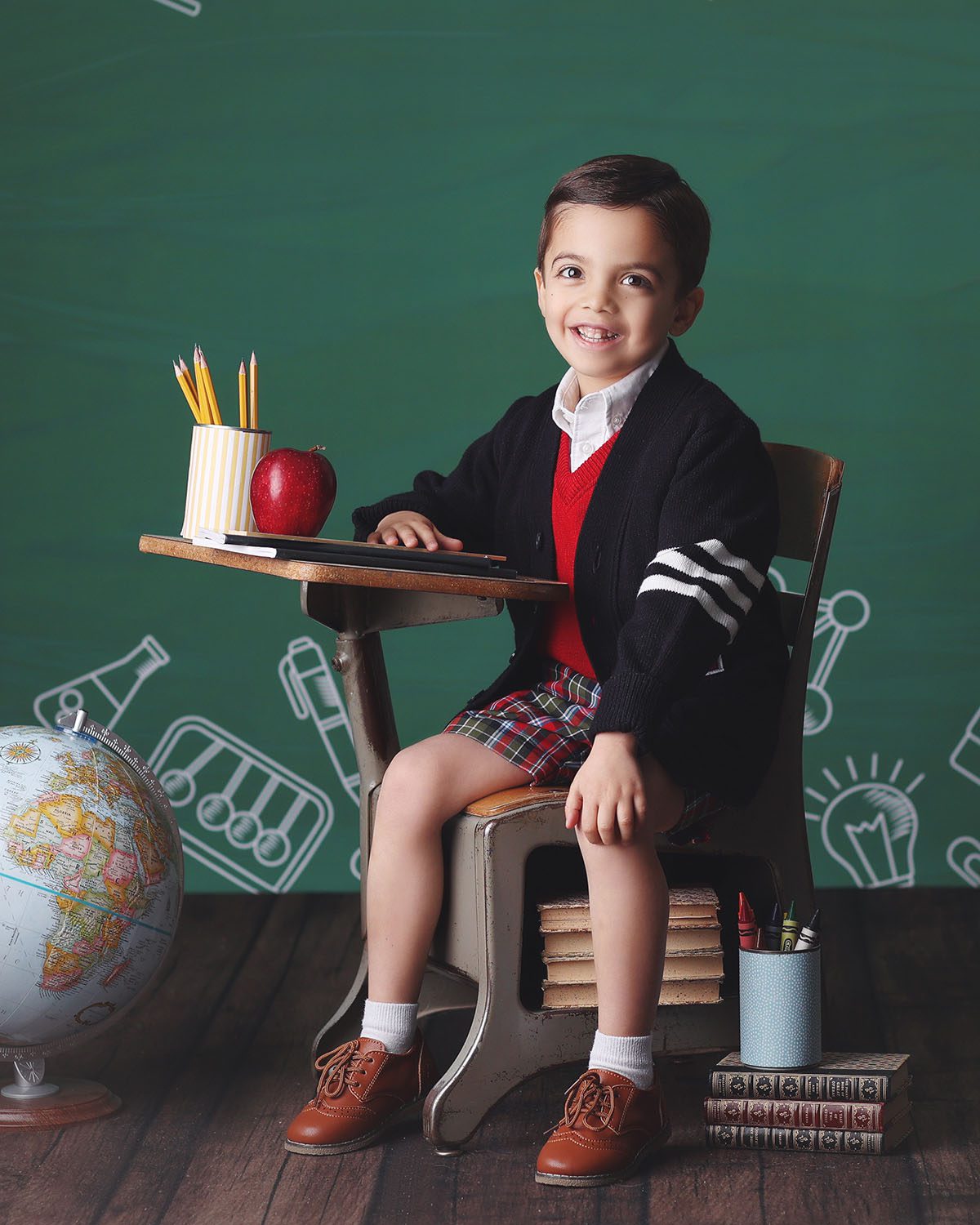 Studio back to school portrait of a young boy wearing a letterman sweater and plaid shorts sitting at vintage school desk with an apple and a globe against a chalkboard backdrop