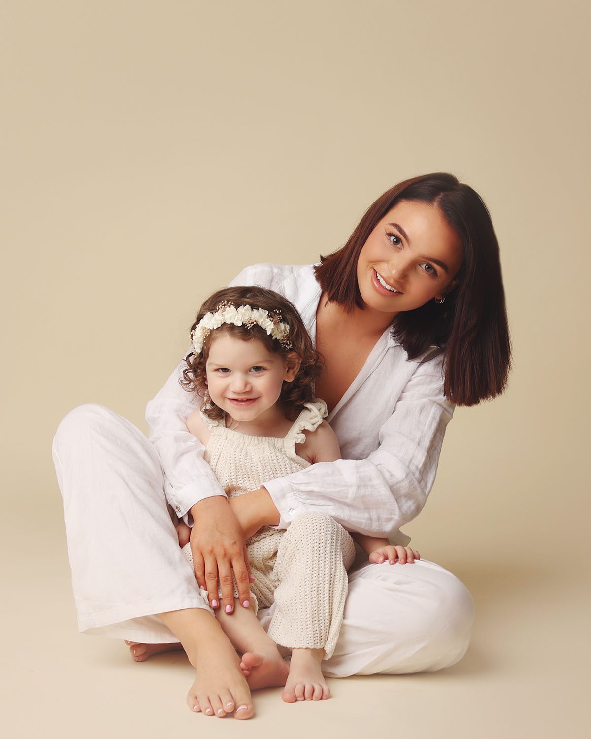 Studio portrait of a mother dressed in white sitting barefoot on a cream backdrop with her daughter in a knit romper smiling at the camera