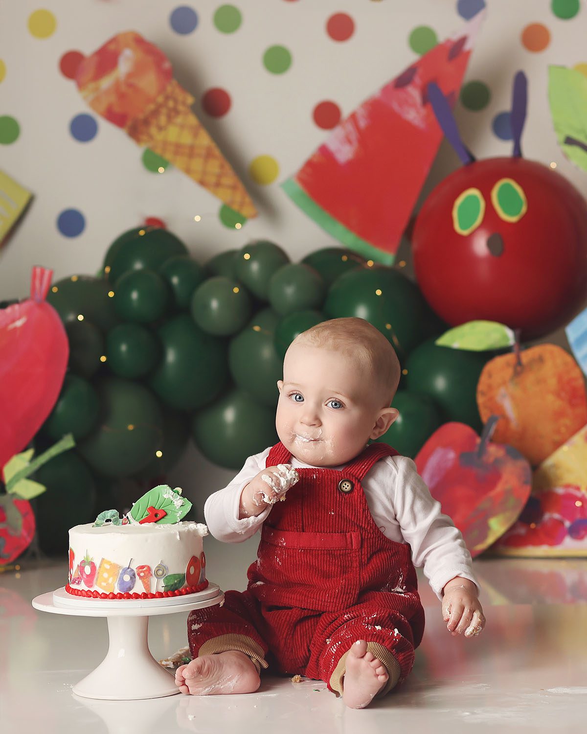 Studio cake smash portrait of a one year old boy wearing red overalls during a Very Hungry Caterpillar themed cake smash, featuring a balloon caterpillar backdrop, oversized props, and a messy moment as he eats his cake