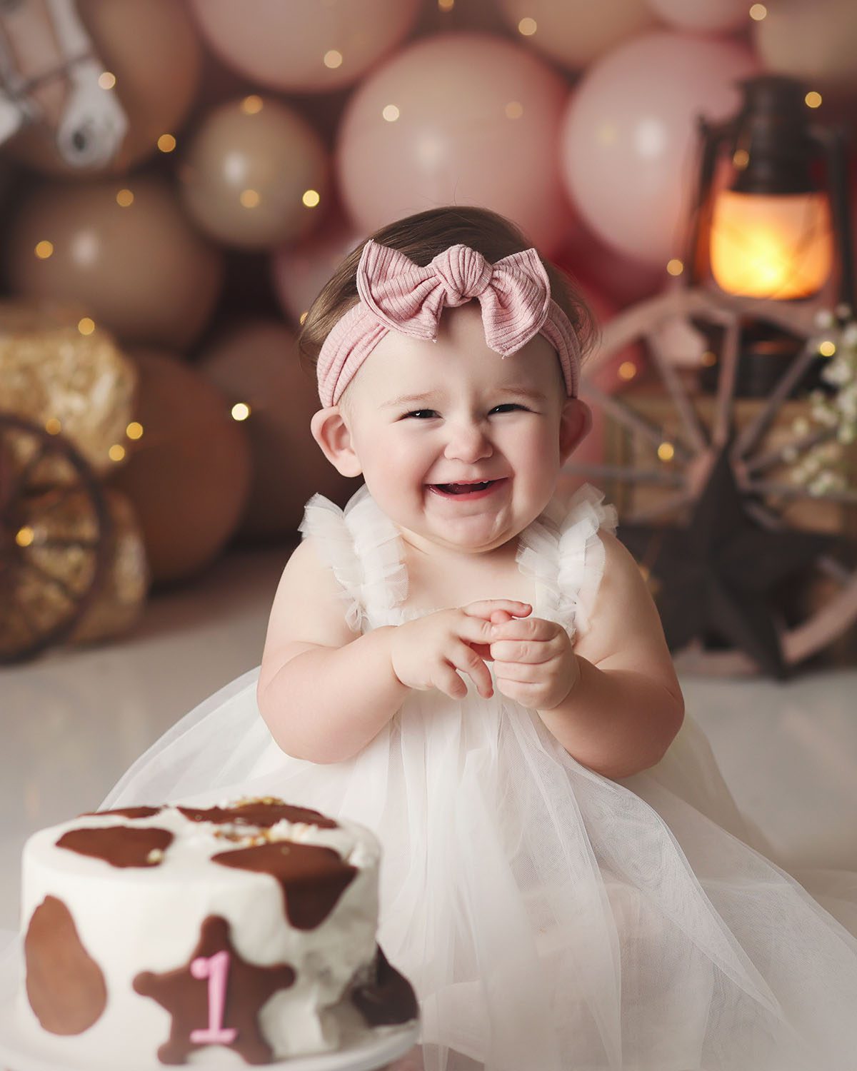 Close up studio cake smash portrait of a one year old girl styled in a white tulle dress with a pink bow during a western themed cake smash, featuring a cow print cake and soft neutral tones.
