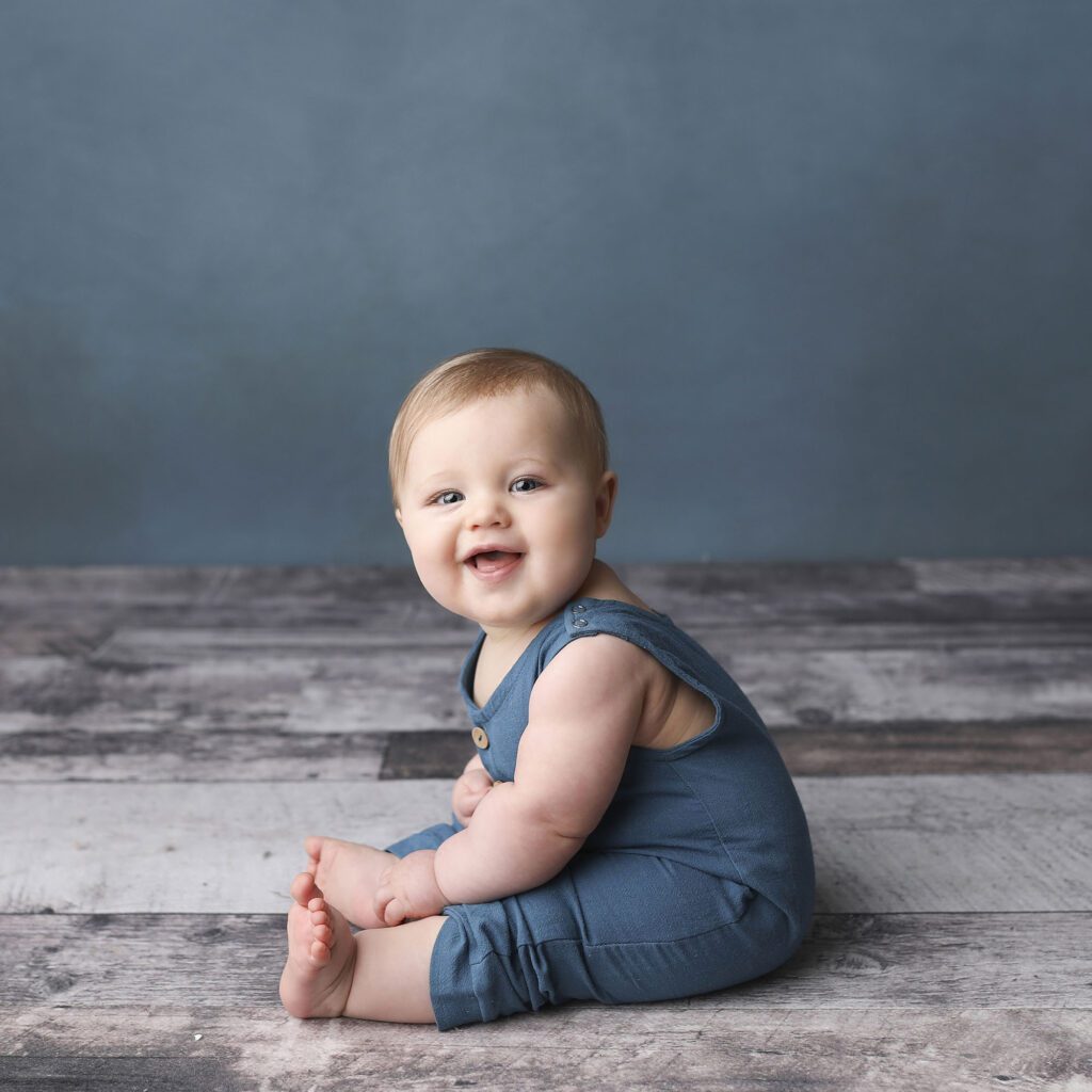 Smiling baby boy wearing blue ovralls during a sitter session on a blue backdrop