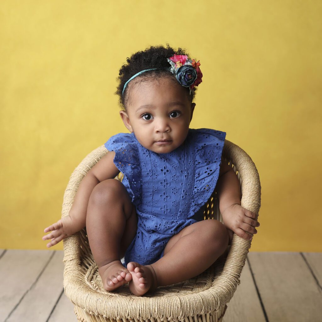Baby girl wearing a blue romper sitting in a small chair with a yellow background