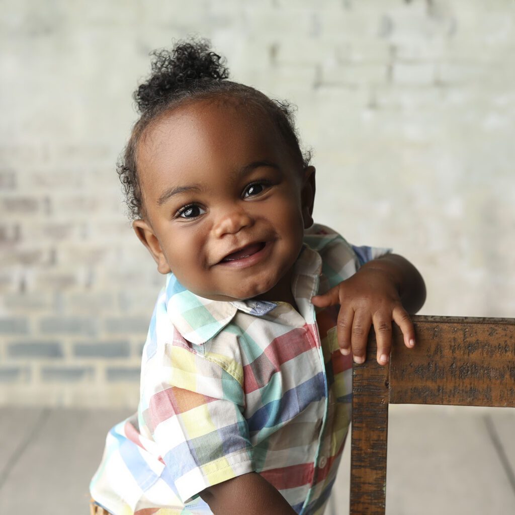 Smiling baby boy holding a chiar while standing in front of a brick background