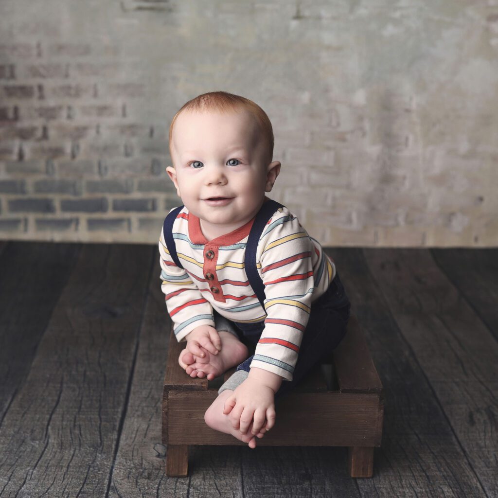 Red haired baby boy sitting on a small bench holding his toes with a brick backdrop
