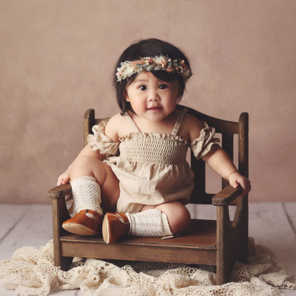 Baby girl sitting on bench wearing a boho outfit and flower crown on a taupe backdrop