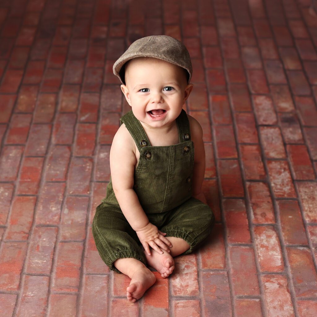 Smiling baby boy wearing green overalls and a brown hat sitting on a brick walkway