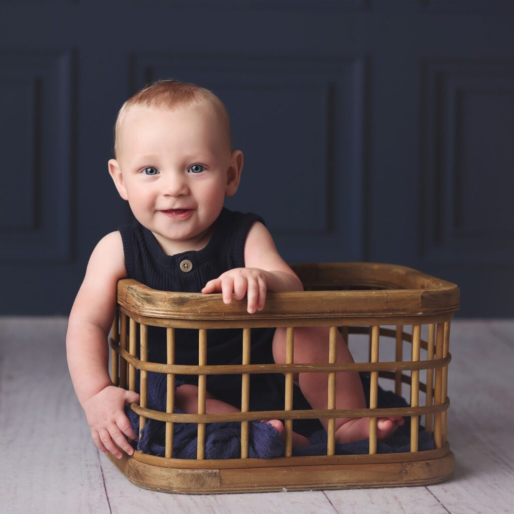 Baby boy sitting inside a wooden crate wearing a blue romper with a navy backdrop