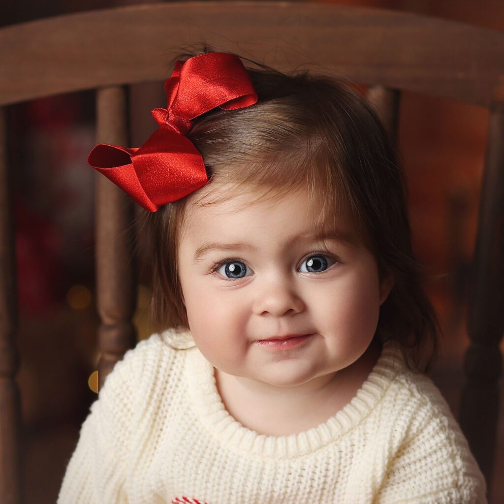 Close up of a smiling baby girl with brown hair and a red bow sitting in a wooden chair