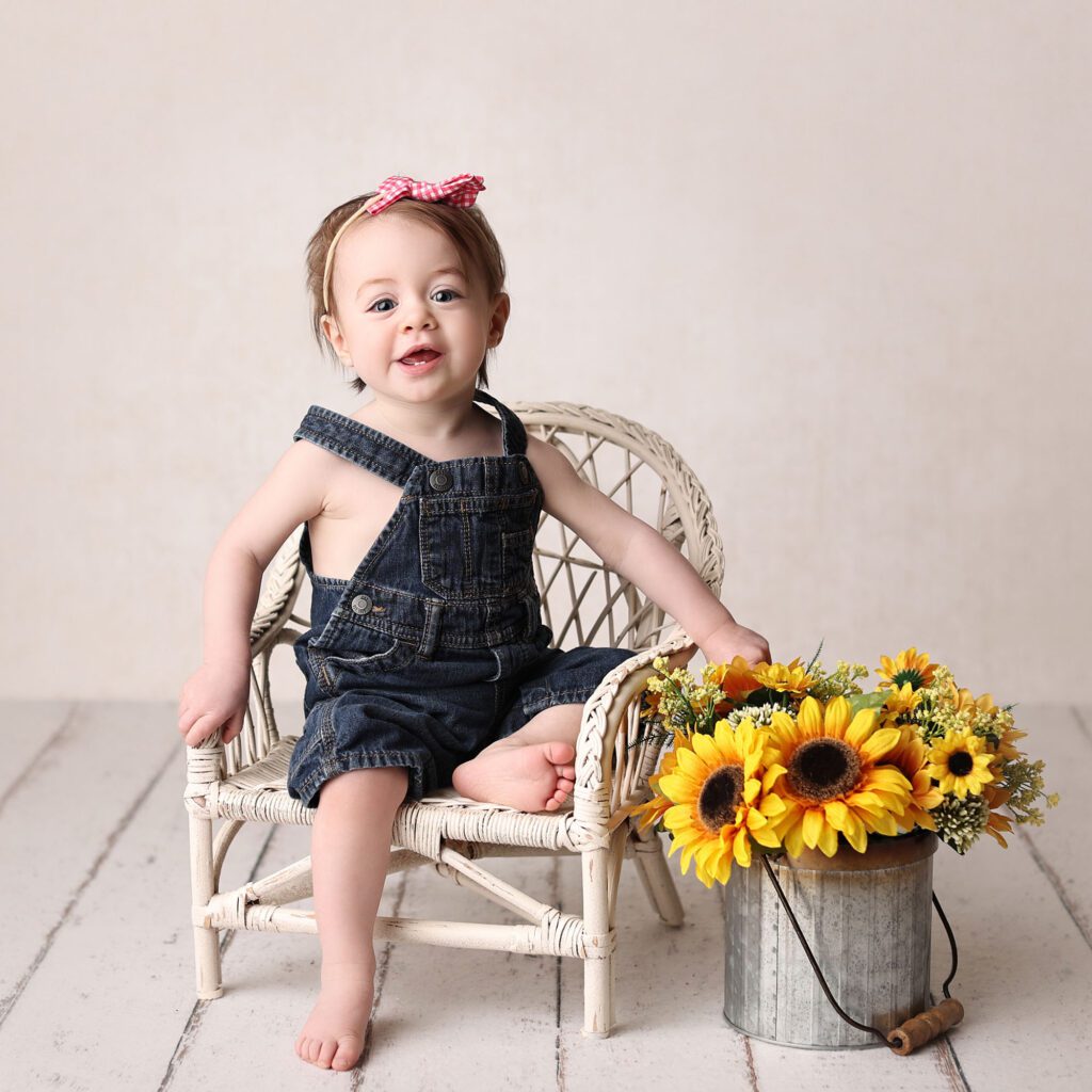 Baby girl wearing jeans and suspenders sitting on a blue backdrop and smiing up at the camera