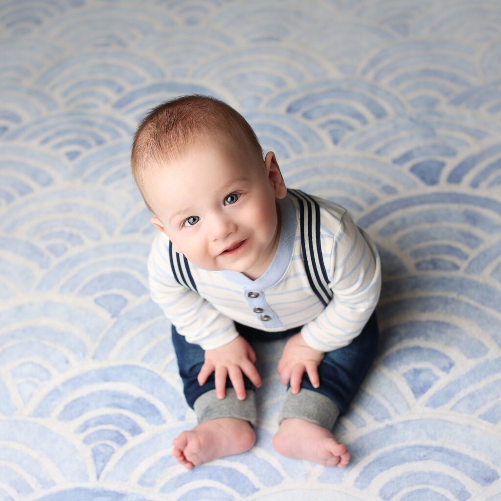 Baby boy wearing jeans and suspenders sitting on a blue backdrop and smiling up at the camera