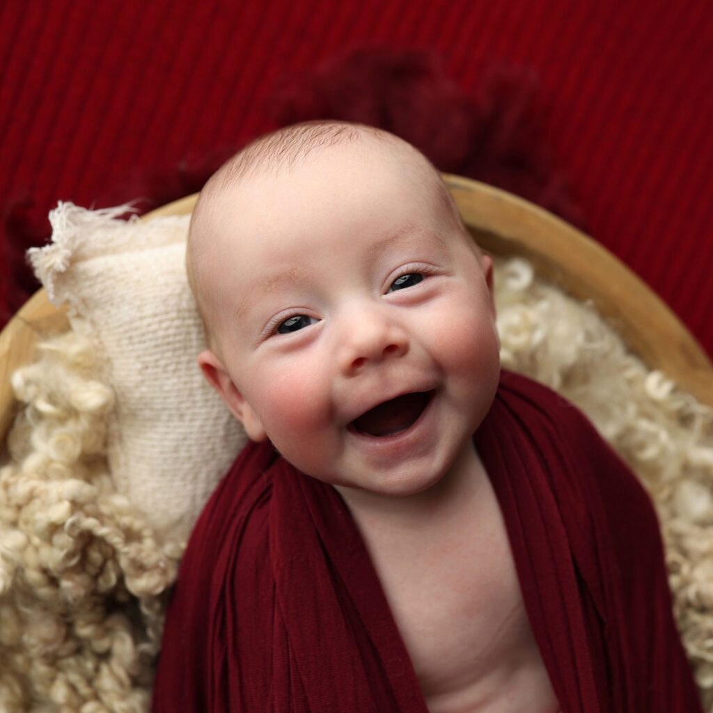 Laughing baby lyiing in a basket wrapped in red fabric and looking at the camera