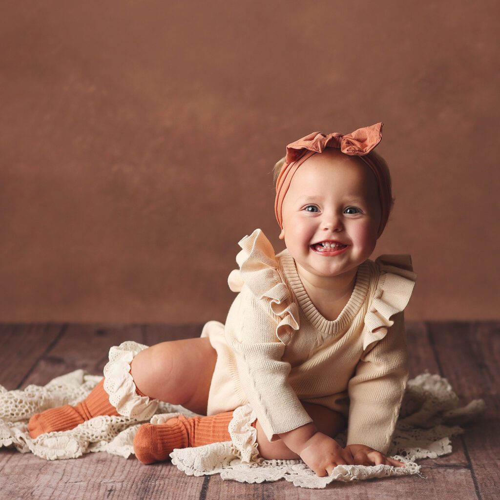 Laughing baby girl wearing a sweater romper and rust knee socks on a brown backdrop