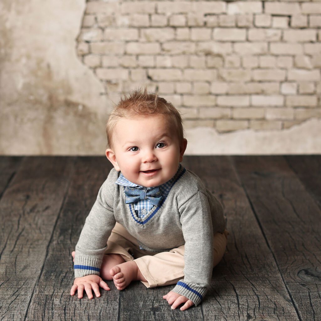 Smiling aby boy wearing a sweater and bow tie sitting in front of a brick backdrop