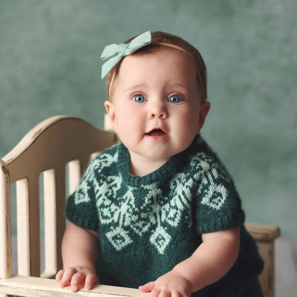 Close up of a baby girl wearing a green sweater sitting on awhite bench with a green backdrop