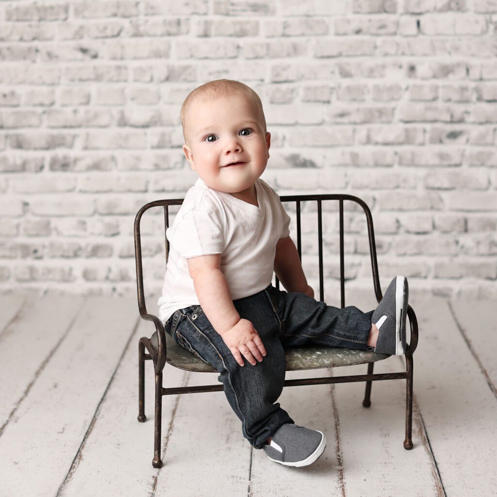Baby boy wearing jeans and a white tshirt sitting on a metal bench in front of a brick backdrop