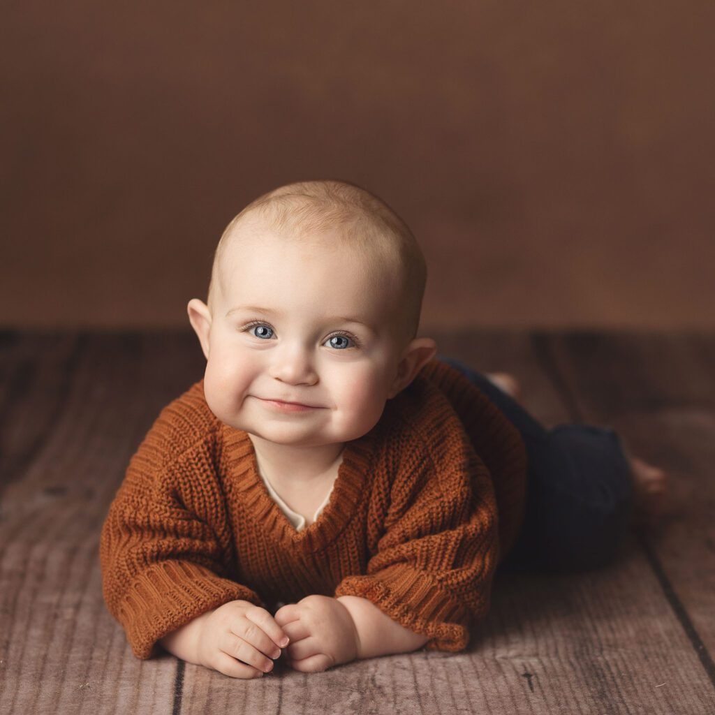 Smiling baby boy wearing a rust colored sweater lying on a wood floor and a brown backdrop