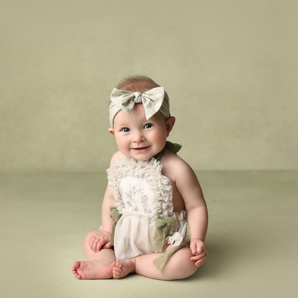 Baby girl wearing a sage green romper and bow sitting on a matching sage green backdrop
