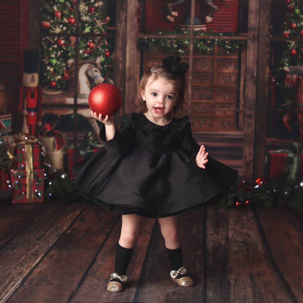 Toddler girl in black dress hilidng a red ornament during a Christmas photo session