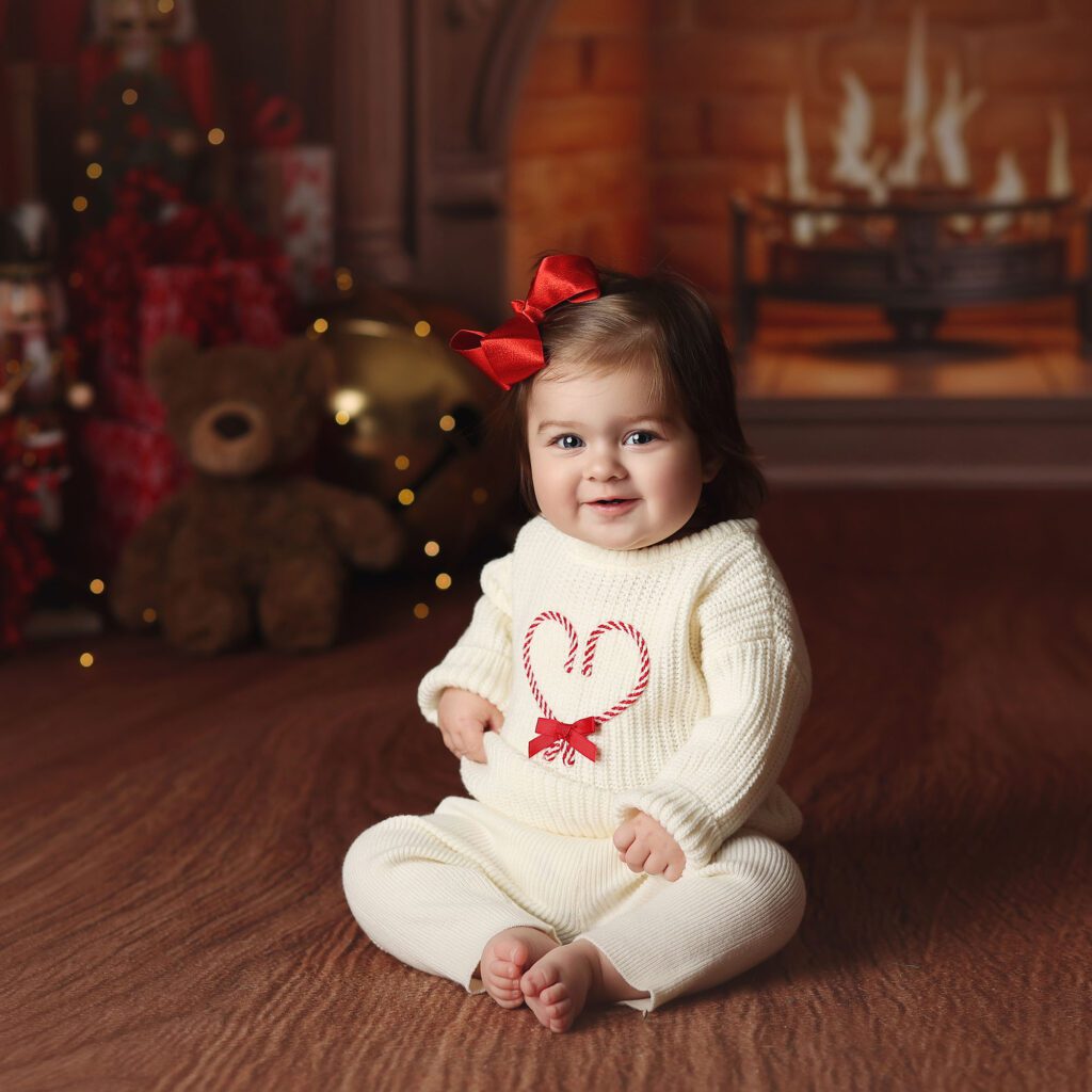 smiling baby girl in a white sweater with candy cane heart sitting by a Christmas fireplace