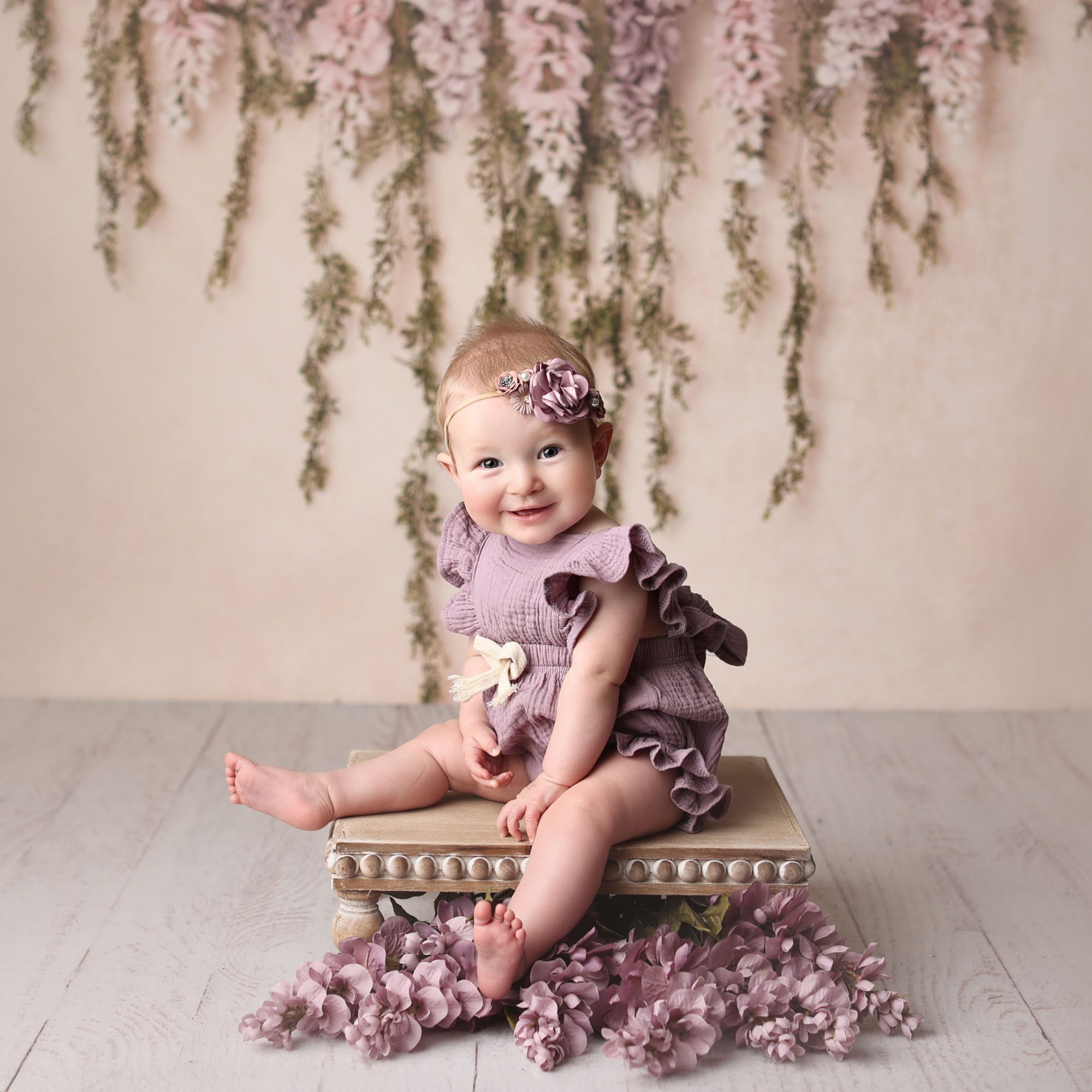 Blue Springs, MO baby girl sitter session in purple romper on wooden platform with a floral garland backdrop