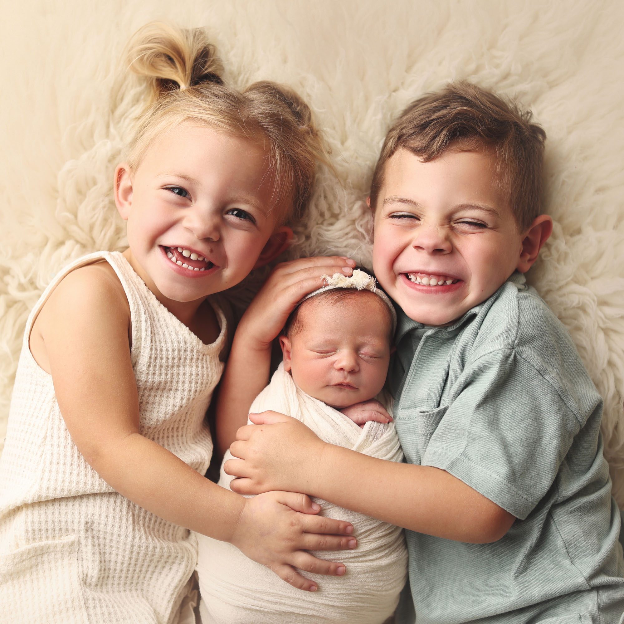 Liberty, MO toddler brother and sister holding newborn baby sister on fur rug smiling at camera