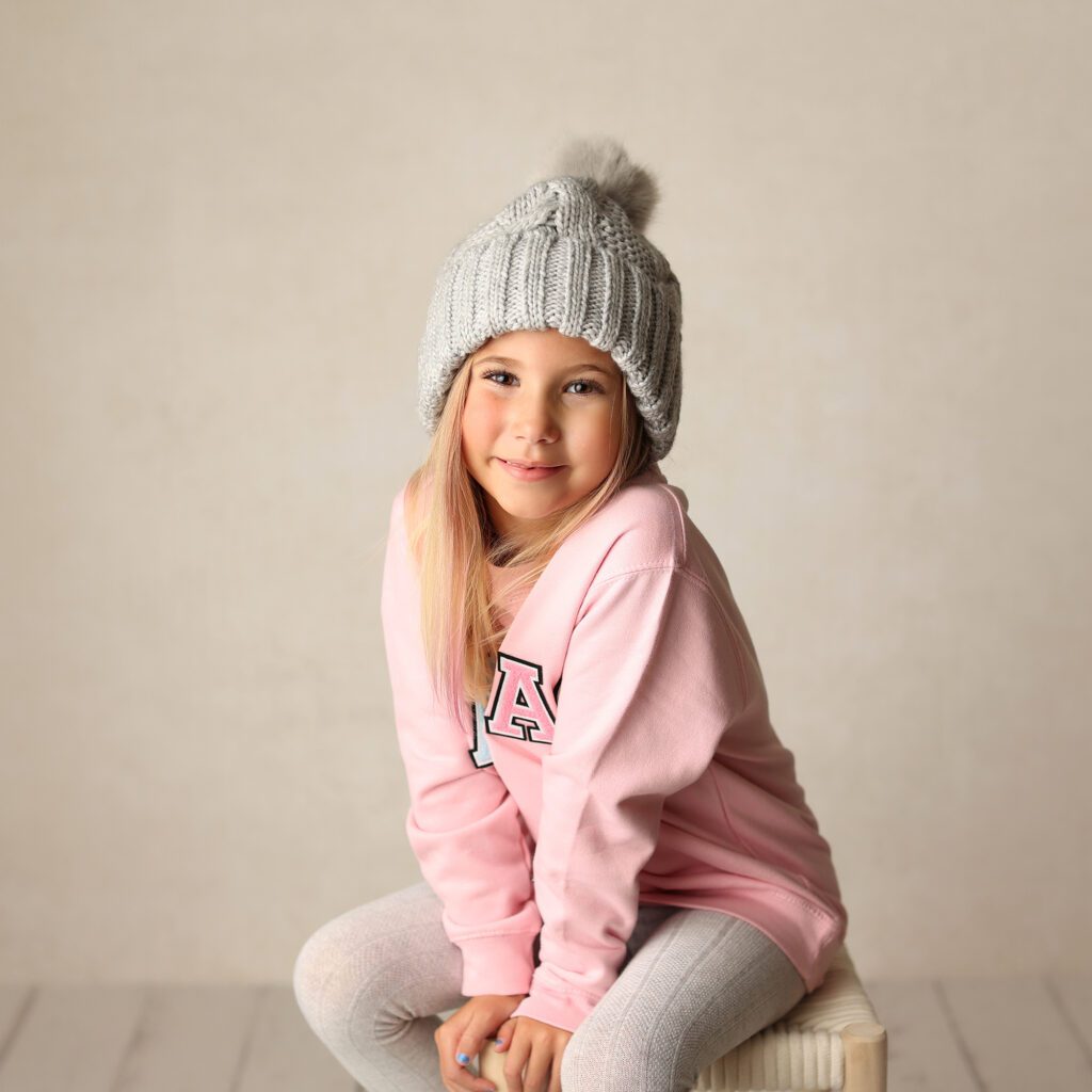 Little girl wearing a gray hat and pink sweatshirt smiling in a studio portrait