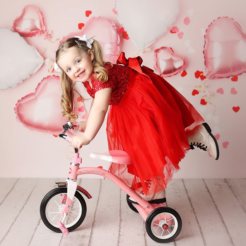 Little girl in a red tulle dress posing ina studio portrait on a pink tricycle
