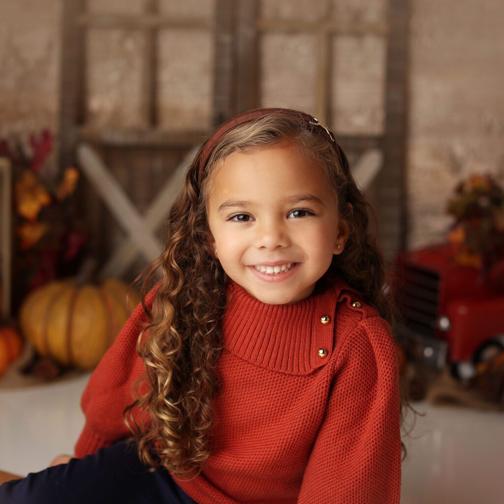 little girl with curly hair wearing an orange sweater in a fall studio portrait