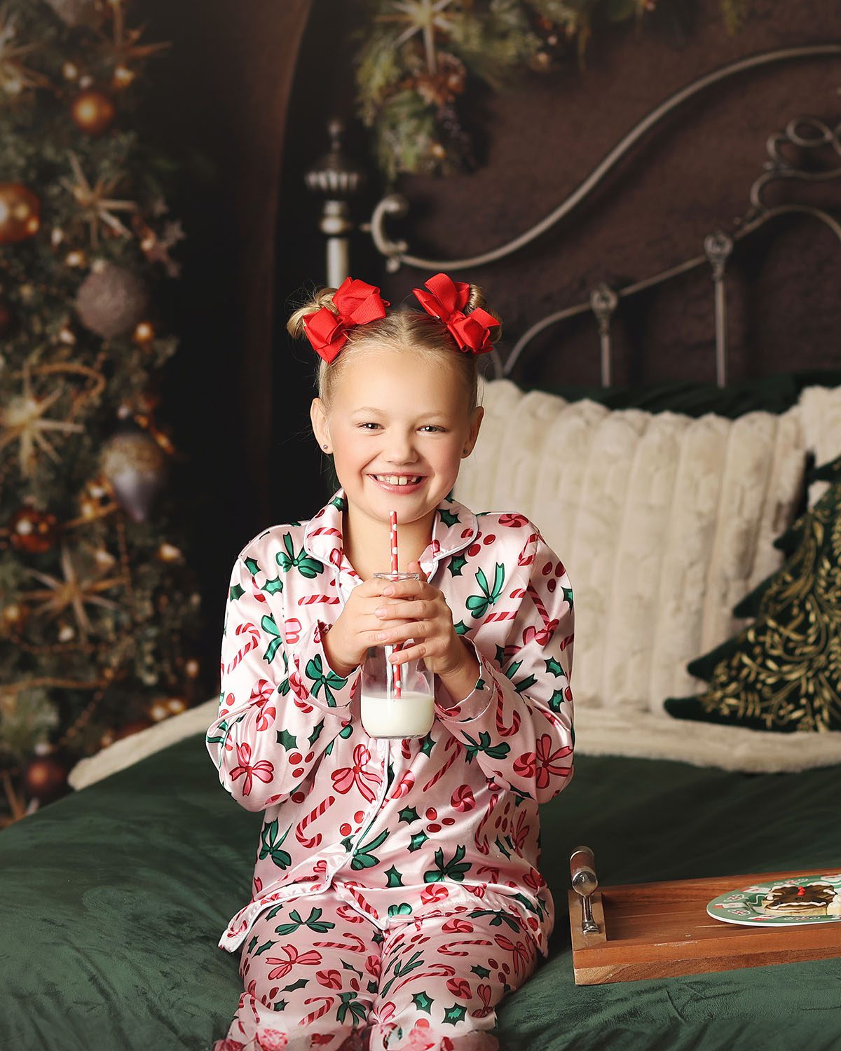 Little girl in red bows and Christmas pajamas sitting on a bed with cookies and milk at her holiday photography session.