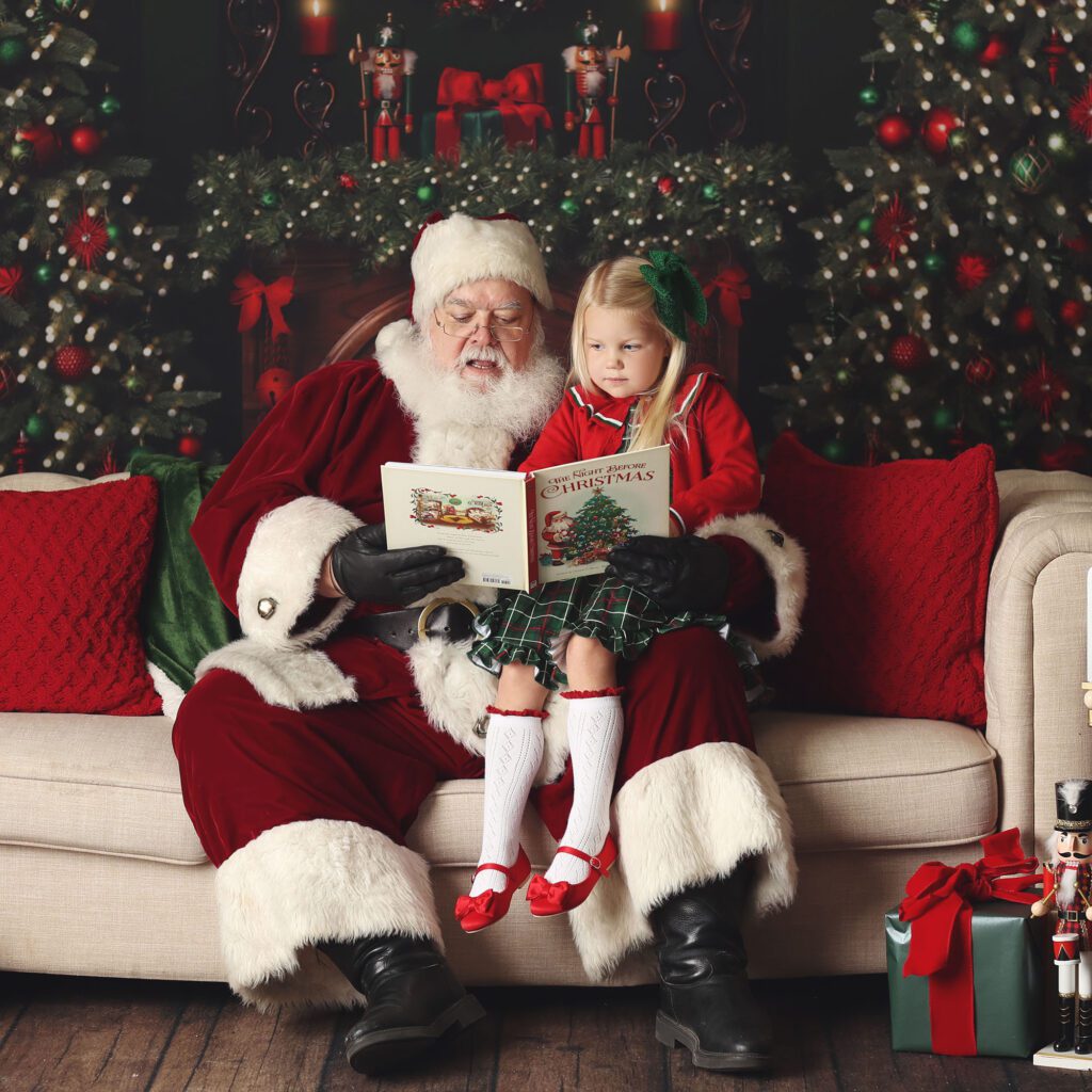 Santa reading a book to a little girl sitting on his lap on a couch