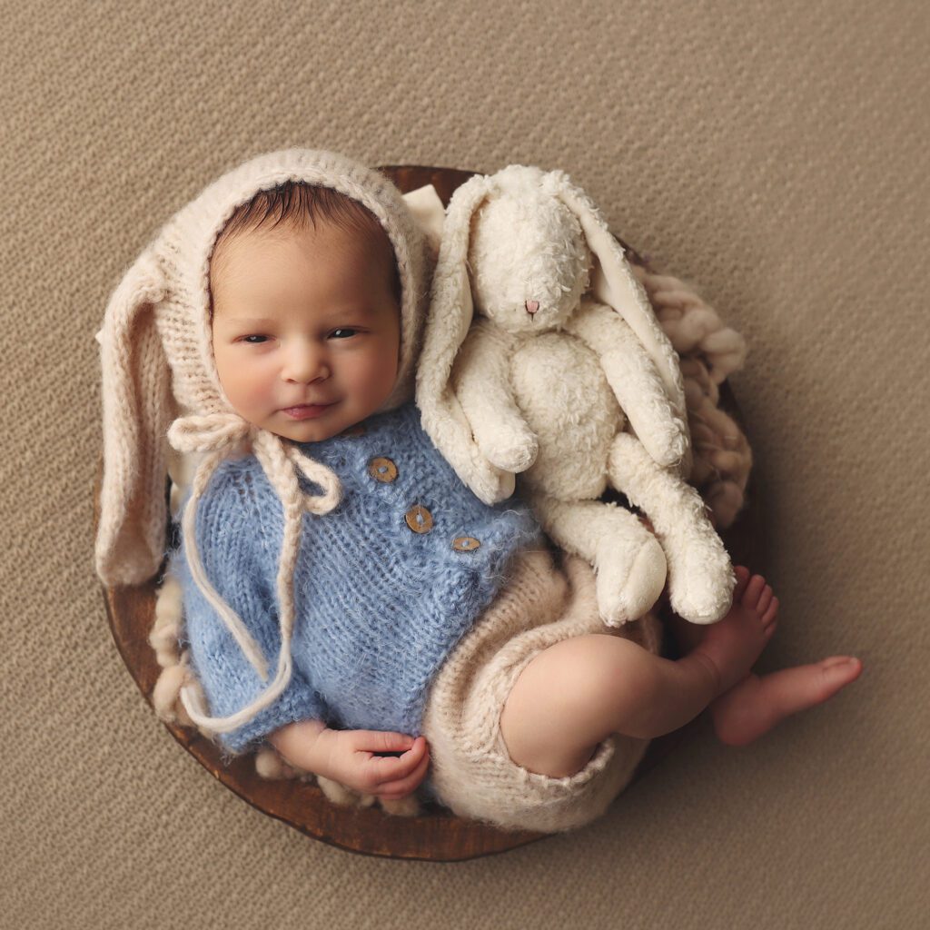 Newborn baby boy awake and smiling while wearing a knit bunny hat and holding a matching bunny
